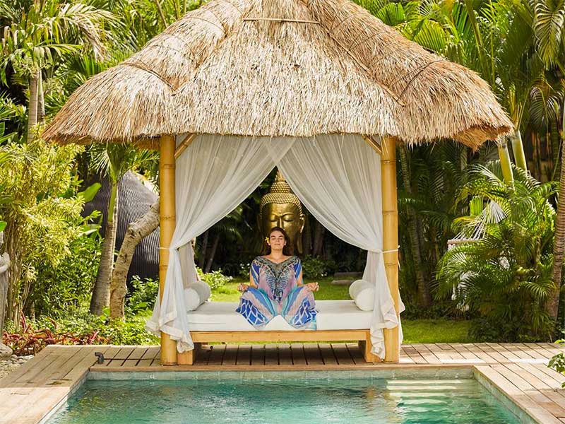 A woman meditates under a small bamboo and grass shelter. It is positioned in front of a swimming pool within a tropical garden.